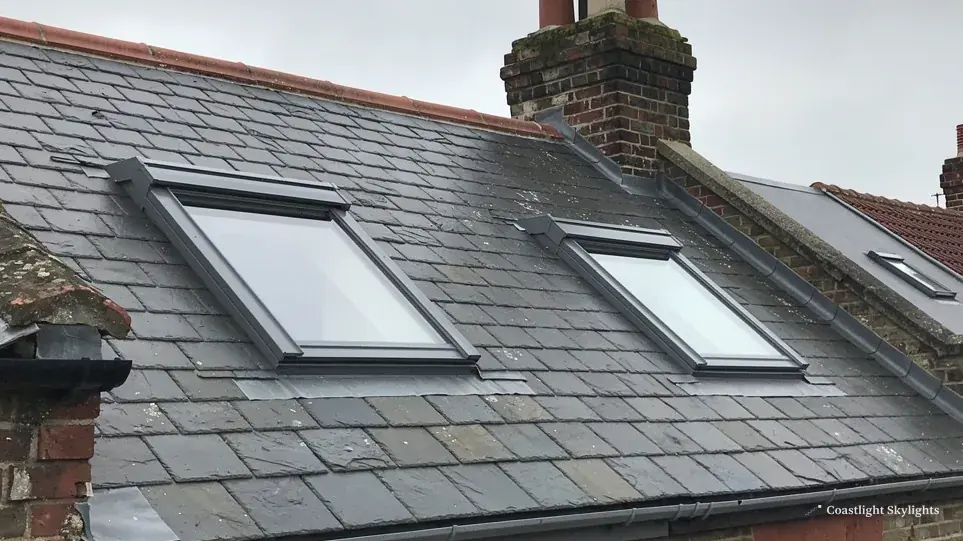 Two roof windows set into a grey slate roof beside a brick chimney