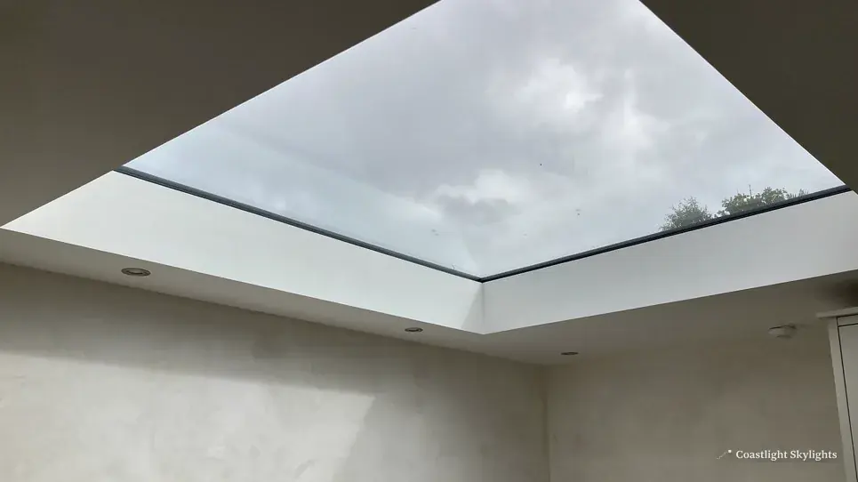 Large flat rooflight in a white ceiling with grey cloud visible through the glass