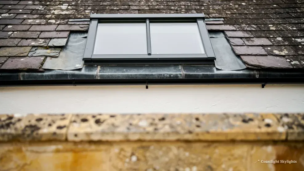 Slim roof window set into a dark slate roof above a stone coping